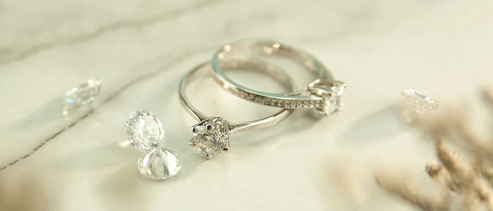 A diamond engagement ring and a band on pale marble beside loose stones and dried flowers