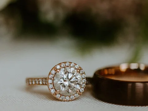 A halo diamond engagement ring in rose gold beside a rose gold band on a pale surface